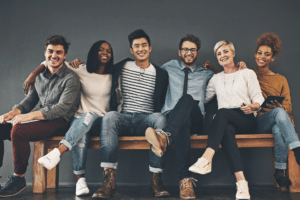 Diverse group of smiling young adults sitting side by side on a long wooden bench against a plain wall